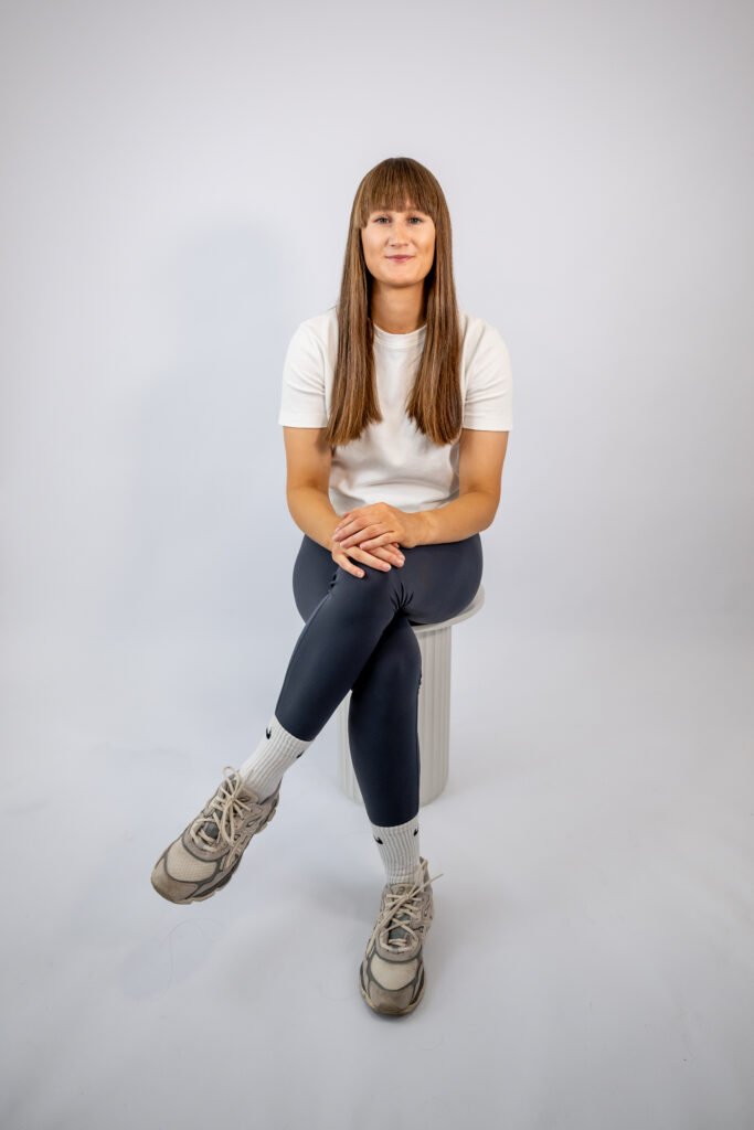 A woman with long brown hair sits cross-legged on a stool against a plain gray studio backdrop, wearing a white shirt, dark leggings, and hiking boots, with a calm, friendly expression.